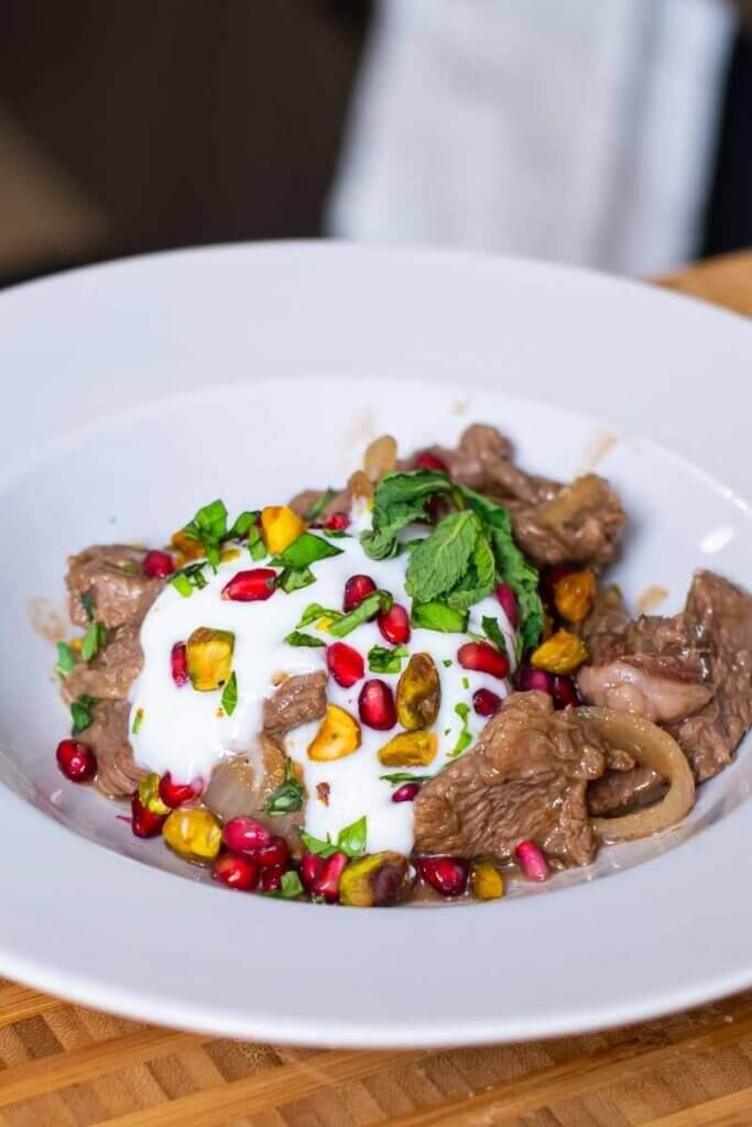 side shot of easy lamb stir fry topped with yogurt, pomegranate seeds, and pistachios in a white serving bowl