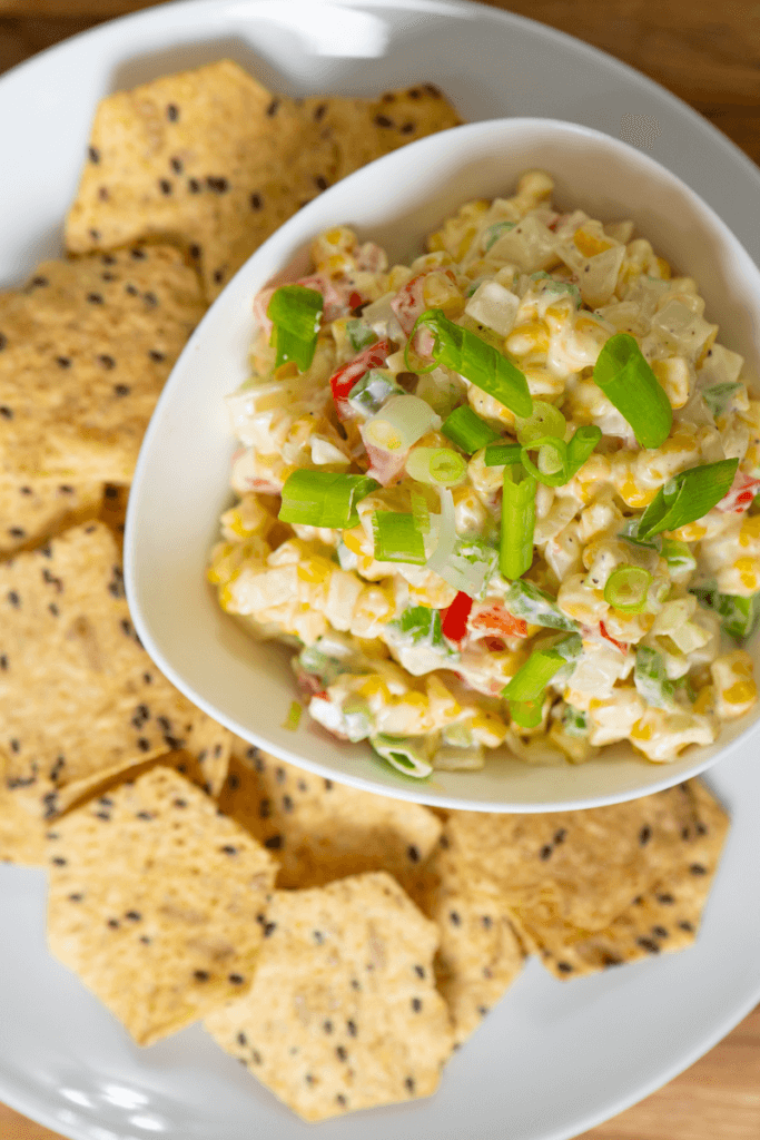 Cheesy corn dip garnished with scallions in a bowl sitting on a plate next to crackers.