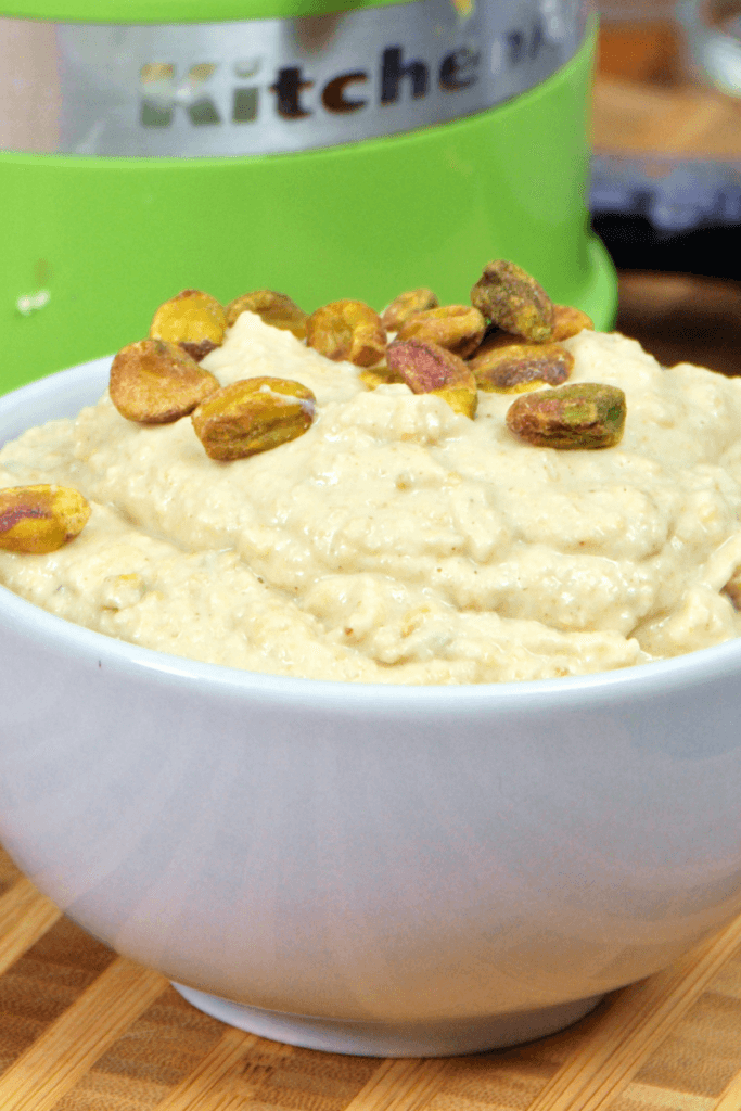side view of pistachio hummus in a white bowl next to a green food processor.