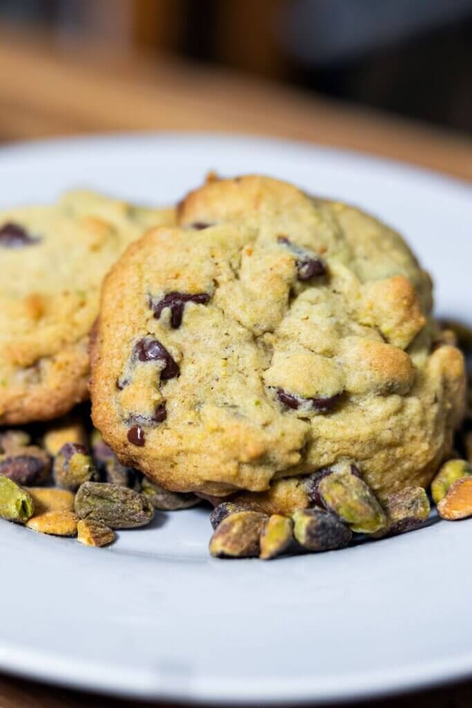 Pistachio chocolate chip cookies on a white plate with shelled pistachios on a plate.